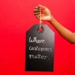 Tangan memegang label bertuliskan "Where customers matter" di latar belakang merah sebagai ilustrasi cara mendapatkan perhatian konsumen